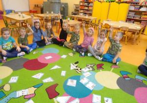 Grupa dzieci siedzi na dywanie z uniesioną ręką wokół rozłożonych na dywanie pociętych na kawałki obrazków ilustrujących zasady z kodeksu dobrych relacji.
